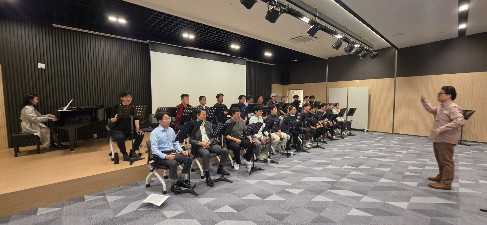 연습홀에서 의자에 나란히 앉은 단원들 앞에서 지휘하는 지휘자, 왼쪽에 피아노 반주자